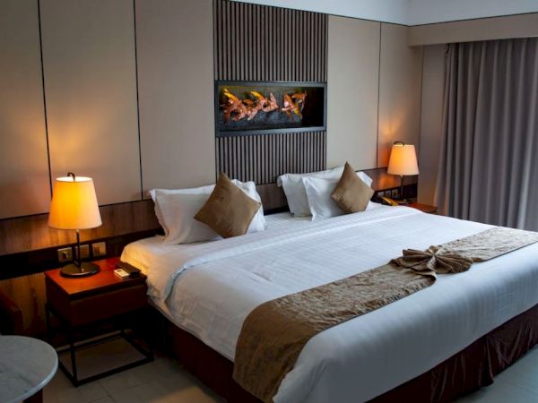 A hotel room features a large bed with pillows, side lamps, a decorative wall element, and curtains by a window, creating a cozy atmosphere.