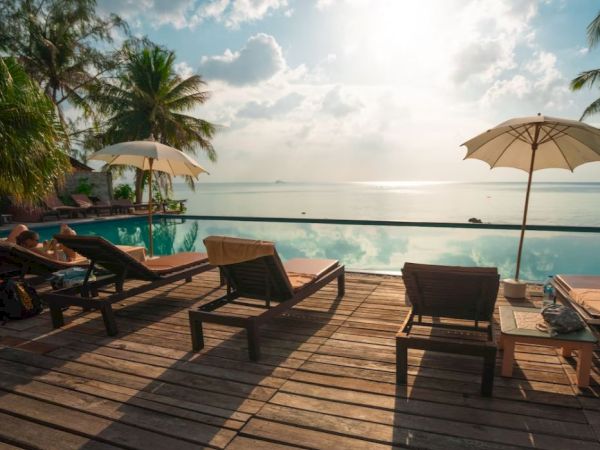 A tranquil poolside scene with lounge chairs, umbrellas, and palm trees, overlooking a calm body of water under a sunny sky.