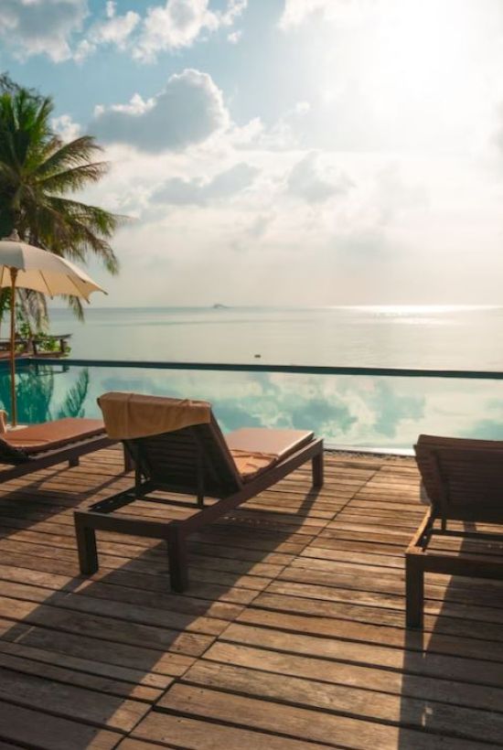Poolside deck with lounge chairs, umbrellas, palm trees, and a view of the ocean under a sunny sky.