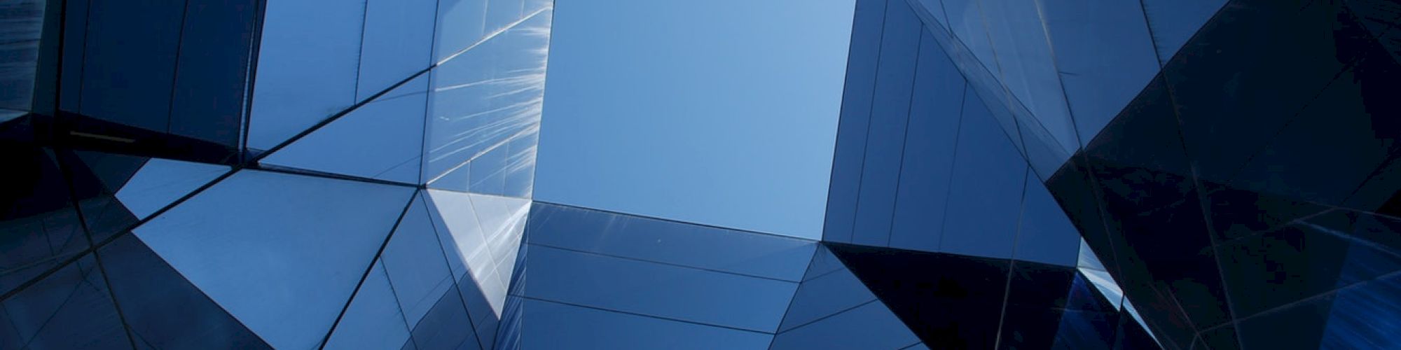 A view looking up through modern, angular glass architecture, creating abstract reflections with a patch of blue sky in the center.