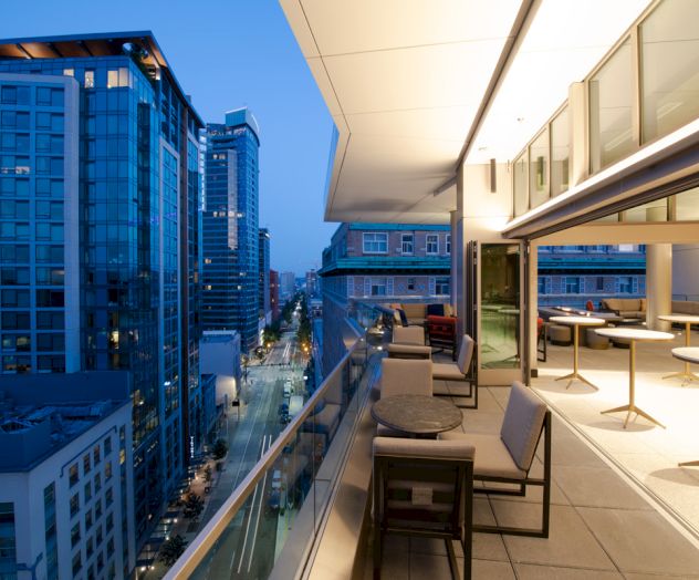 A modern outdoor balcony with tables and chairs overlooks a city street lined with high-rise buildings at dusk.
