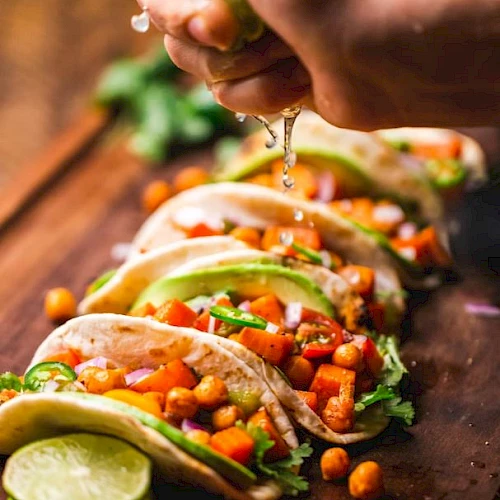 A person is squeezing lime juice over tacos filled with chickpeas, vegetables, and avocado slices on a wooden board.