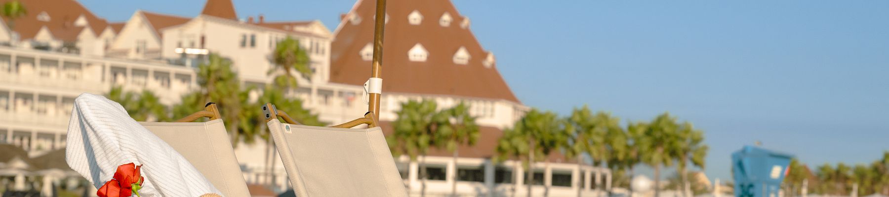 A sunny beach scene with reclining chairs, a large patterned parasol, a red rose, and distant resort buildings.