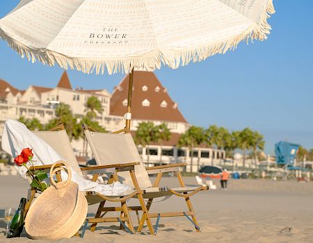 A sunny beach scene with reclining chairs, a large patterned parasol, a red rose, and distant resort buildings.