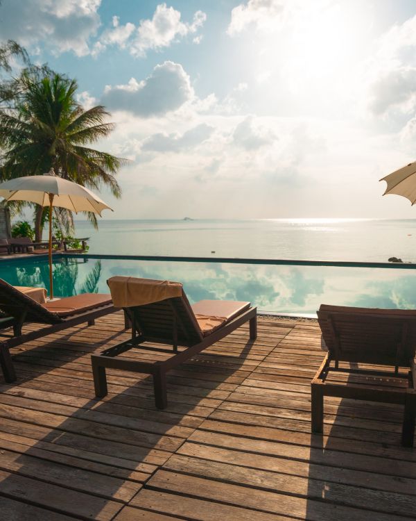 Beachside pool area with lounge chairs, umbrellas, and palm trees overlooking a tranquil ocean under a bright, cloudy sky.
