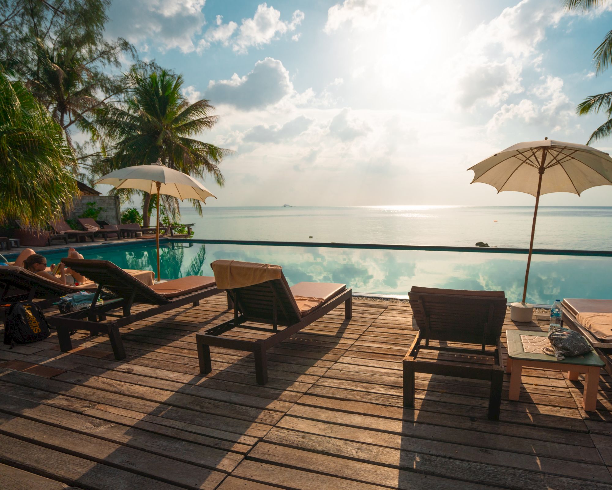 A serene beach setting with lounge chairs, umbrellas, and palm trees by an infinity pool under a cloudy sky.