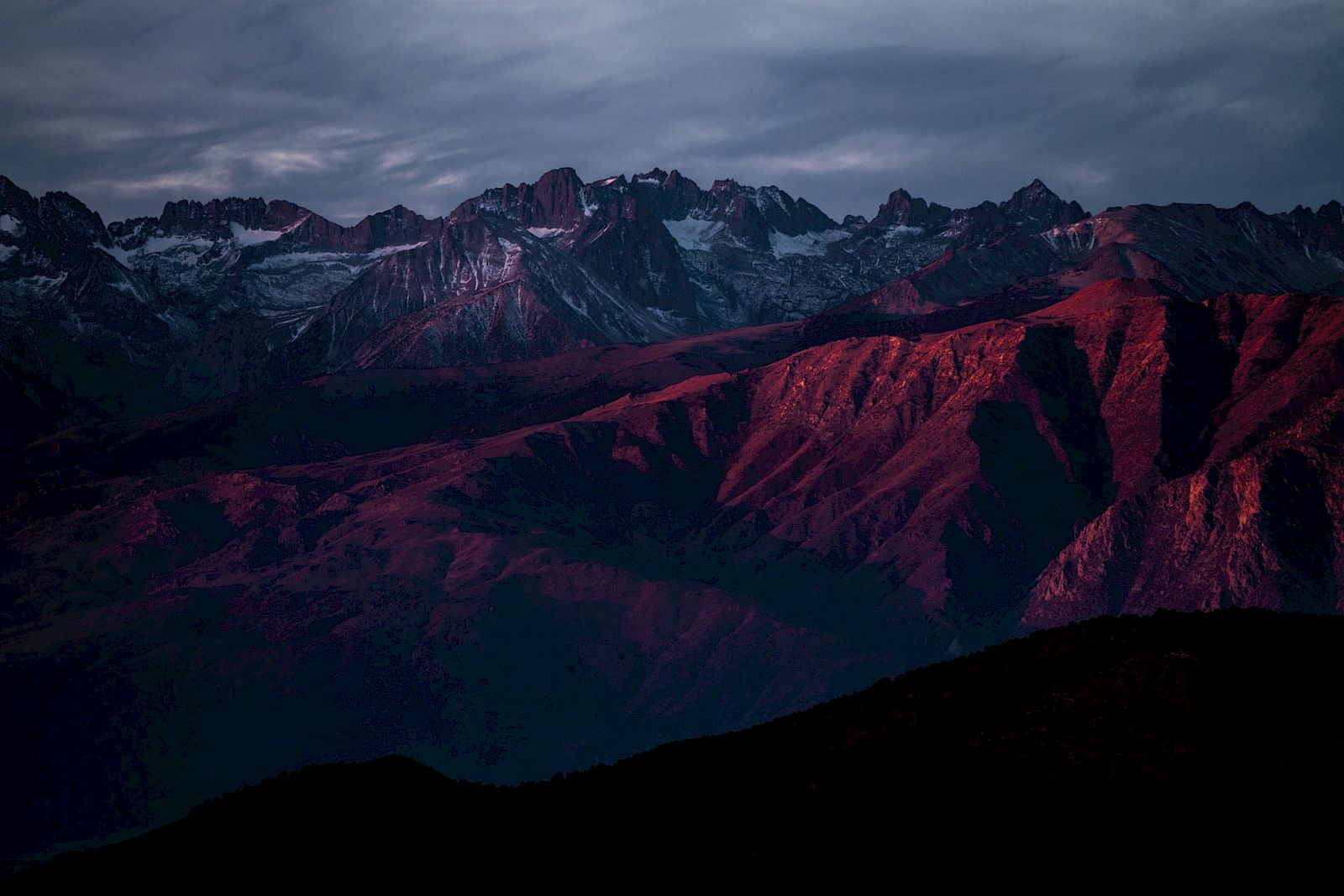 A mountain range at dusk with purplish hues and a dramatic sky, casting red light on rugged peaks.