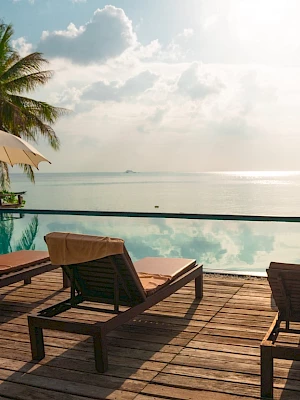Sunlit beach scene with lounge chairs, umbrellas, a pool, and palm trees overlooking the ocean.