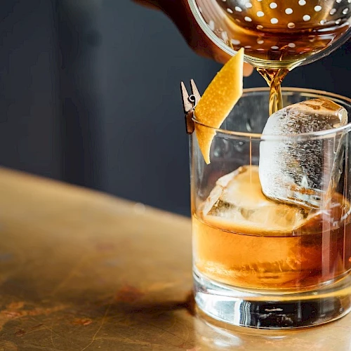 A hand is pouring a brown drink into a glass with ice and an orange peel garnish, set on a table.