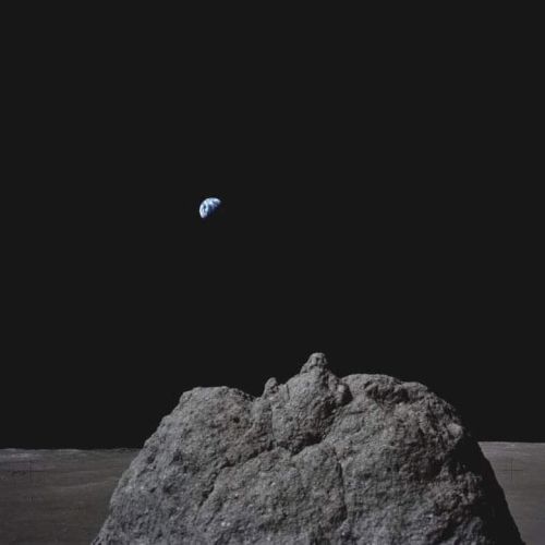 A view of Earth rising over the lunar horizon, with the Moon's surface and a large rock in the foreground.