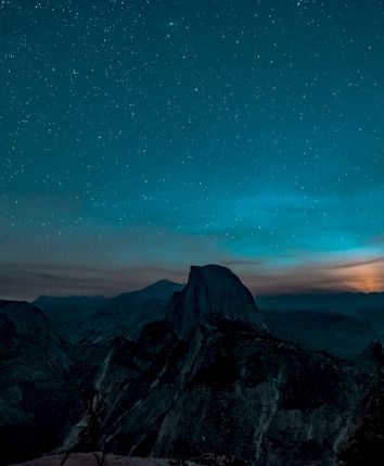 A mountain landscape under a starry night sky, with a glowing horizon and silhouettes of trees in the foreground.