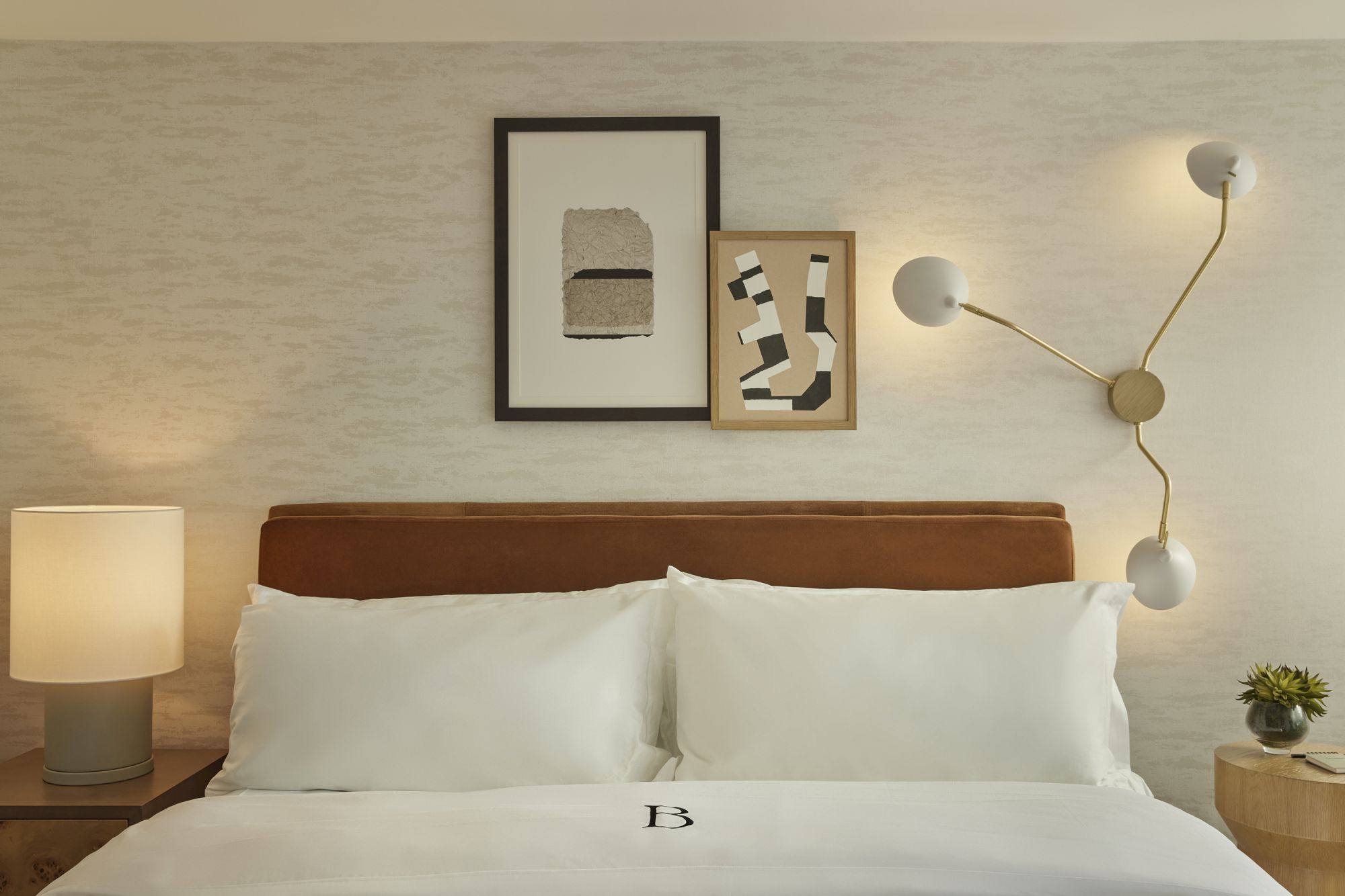 A neatly made hotel bed with white linens, two pillows, a wooden headboard, wall art, a modern wall lamp, and a bedside table with a small plant.