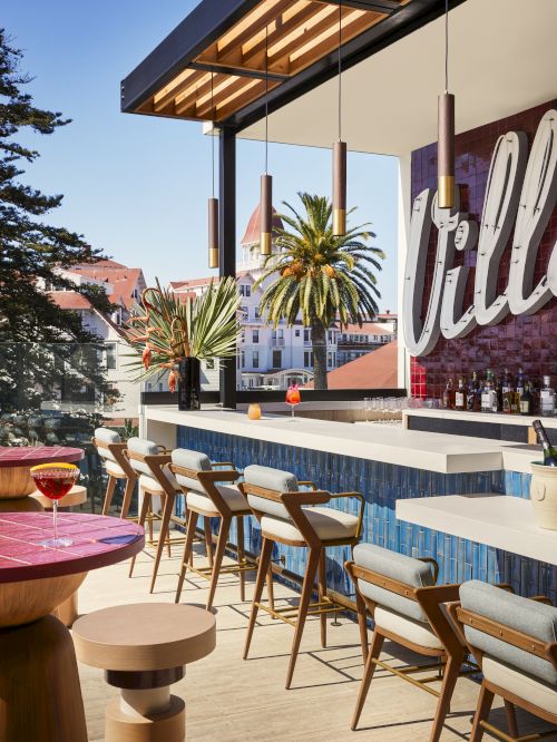 The image shows a stylish outdoor bar with a row of wooden chairs, modern tables, palm trees, and a scenic view in the background.