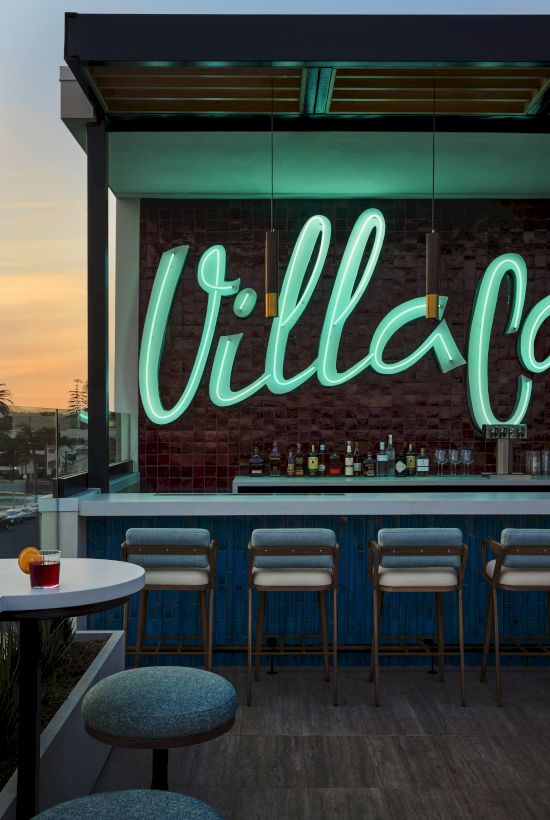 Outdoor bar with a neon sign, stools, and a table with a drink, set against a sunset backdrop.