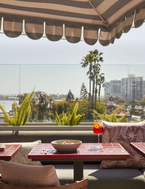 A cozy outdoor dining setup with a striped awning, scenic city view, red table, chairs, and plated meal.
