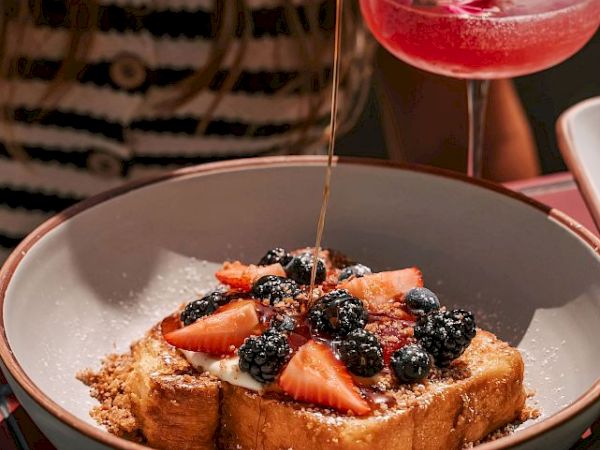A bowl of French toast topped with strawberries and blackberries. A pink cocktail with a flower float beside it on the table.