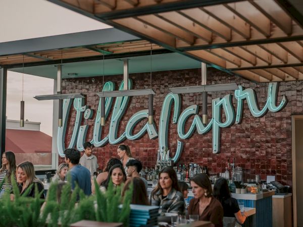 A group of people are socializing at an outdoor venue with a sign that reads "Villa Capri," surrounded by a modern and stylish decor.