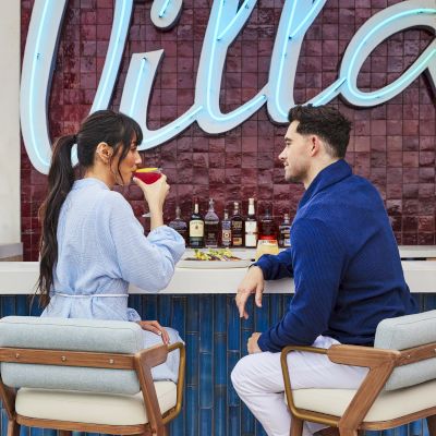 A couple is seated at an outdoor bar, enjoying drinks. A neon sign reading "Villa" is visible in the background, adding ambiance.