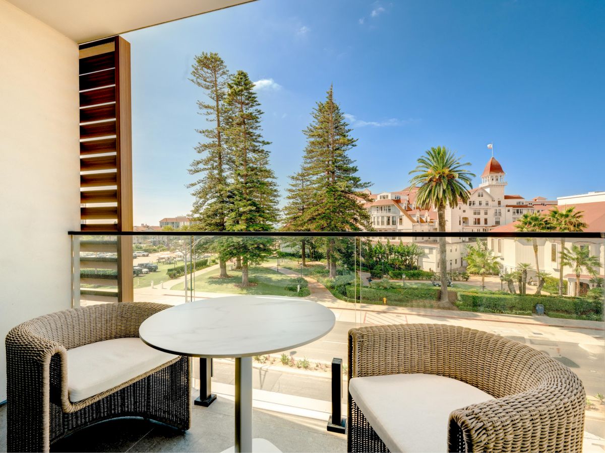 A balcony with two curved wicker chairs and a round table overlooks a sunny plaza with palm trees and a red-roofed building in the distance.