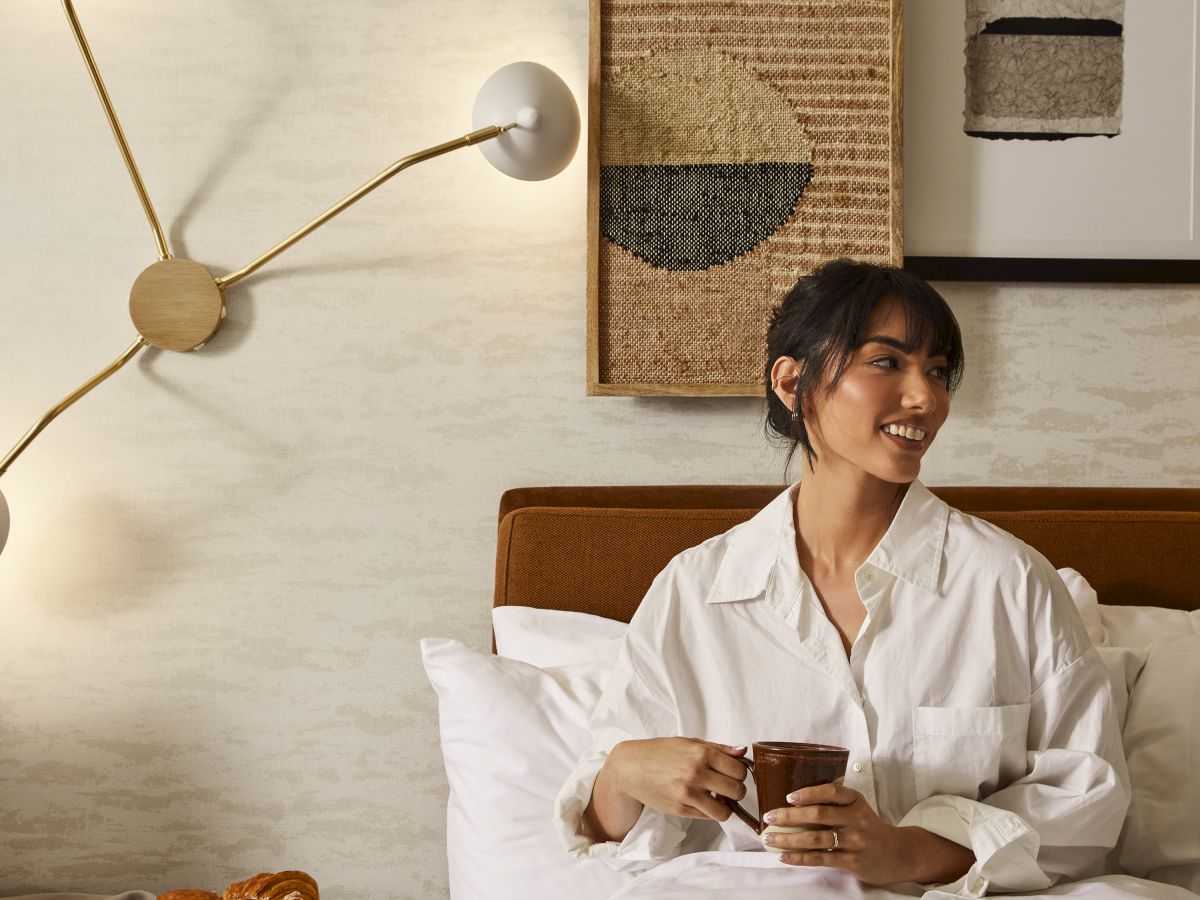 A person sits in bed holding a mug, with pastries on a side table and modern wall art and lighting in the background.