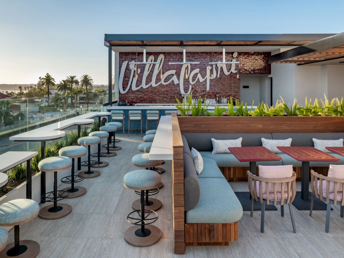 The image shows a modern rooftop restaurant with seating, tables, and a sign reading "Villa Capri," offering a scenic outdoor view.