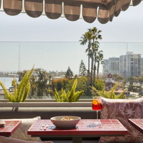 Outdoor dining setup with a table, bowl, drink, and a striped awning. Background features cityscape, palm trees, and plants.