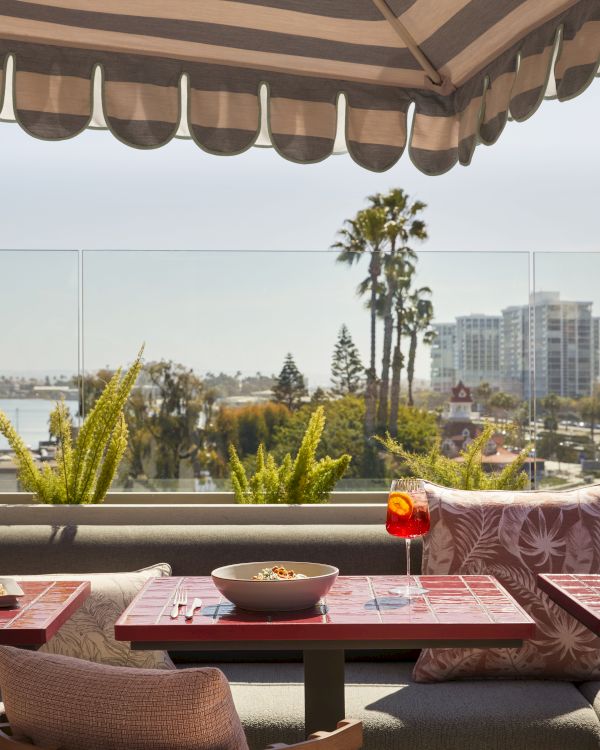 Outdoor dining setup with a table, bowl, drink, and a striped awning. Background features cityscape, palm trees, and plants.
