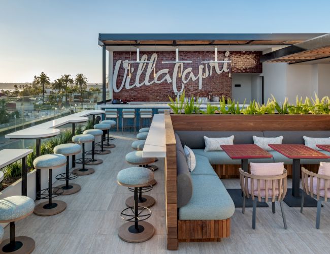 The image shows a stylish outdoor rooftop seating area with tables, chairs, and a sign on a brick wall.