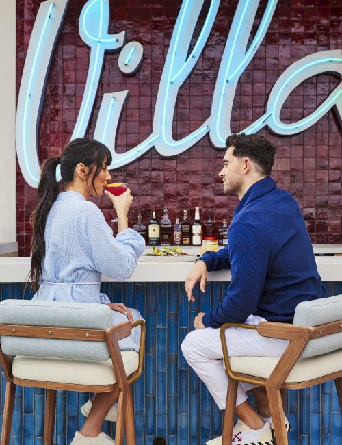 A couple is sitting at an outdoor bar with a "Villa" sign, enjoying drinks and chatting on bar stools.