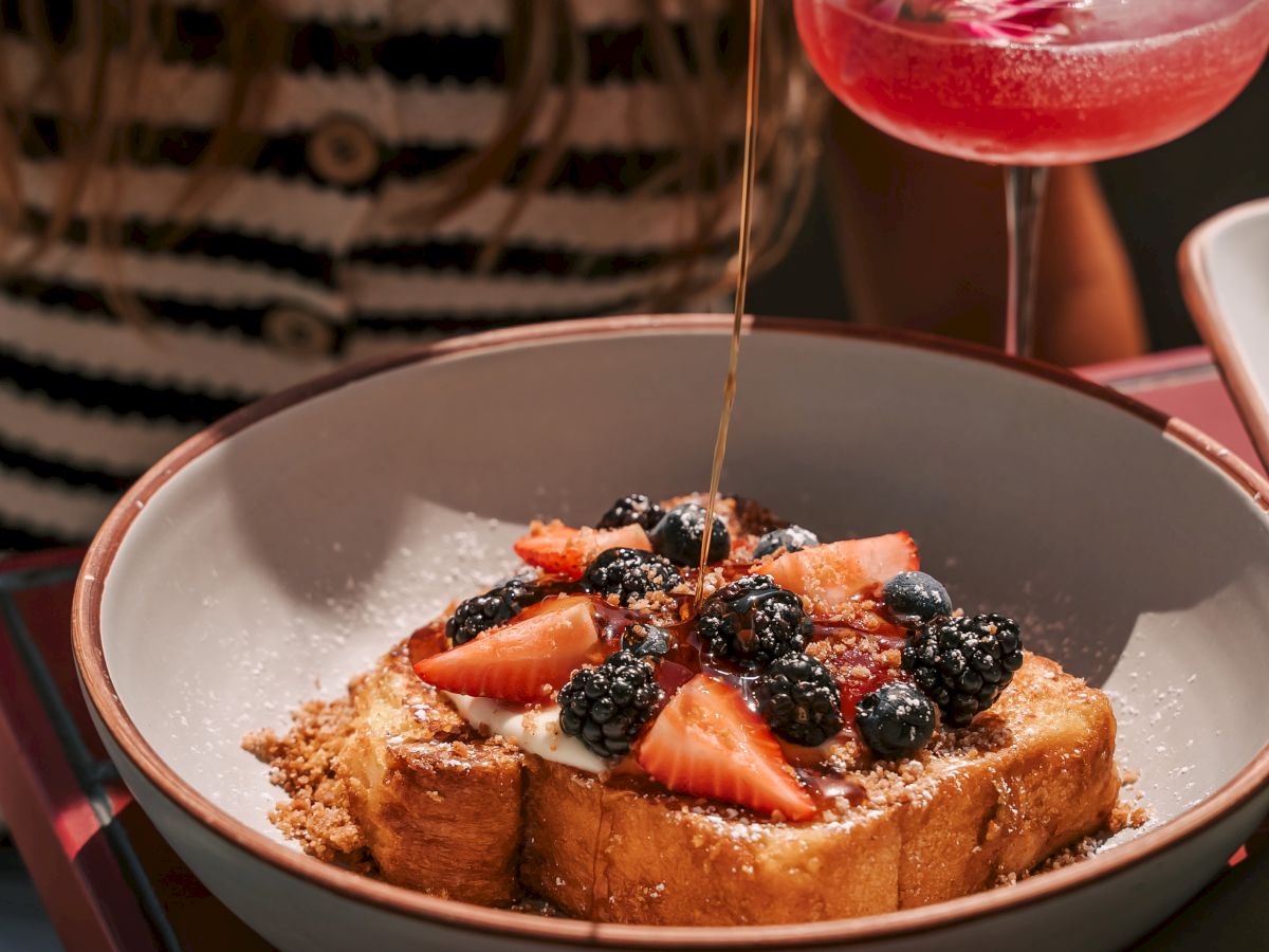A bowl of toast topped with berries next to a pink cocktail garnished with a flower, on a table.