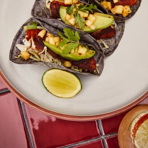 The image shows three tacos with avocado and diced toppings, a lime wedge on the plate, and a lime-topped drink on a red tiled surface.