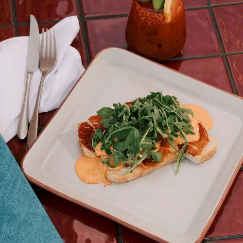 A dish with greens and sauce on toast sits on a square plate, next to a drink, cutlery, and a blue menu on a tiled surface.