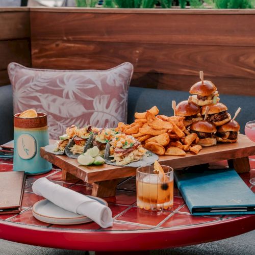 A cozy table setup with sliders, tacos, potato wedges, cocktails, menus, and napkins, surrounded by cushions on a red tiled table.