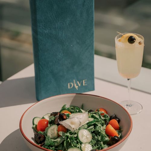 A salad with greens, cucumbers, and tomatoes, a drink with a berry garnish, and a blue menu labeled "DIVE" are on a table.