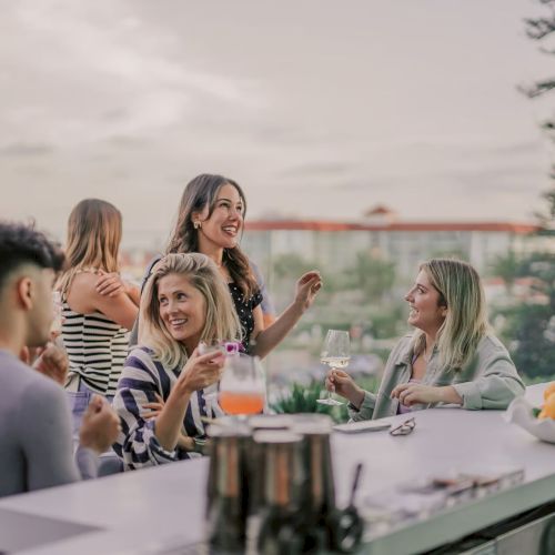 A group of people socializing at an outdoor bar with drinks, surrounded by greenery and a fruit bowl on the counter.