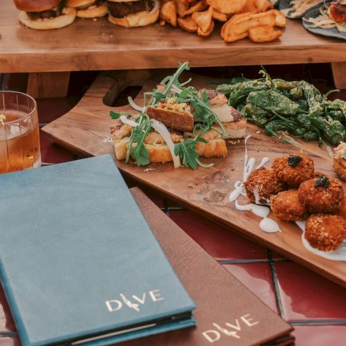 The image shows a table with sliders, fries, appetizers, and drinks. Menus labeled "Dive" are placed in the foreground.