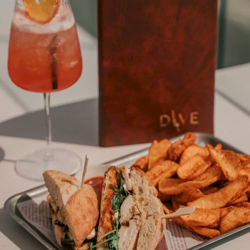 A tray with sandwiches and potato wedges, a red-orange drink with citrus garnish, and a menu standing upright on a table.