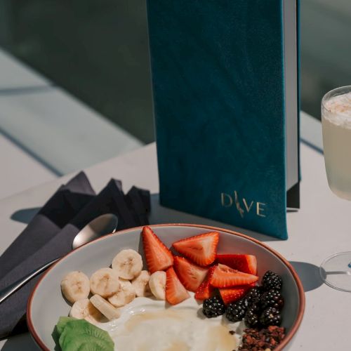 A bowl with yogurt, bananas, strawberries, blackberries, granola, and avocado, next to a blue menu and a drink on a table.