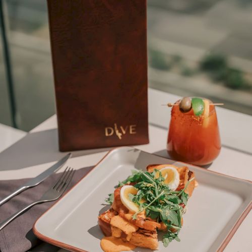 A meal with fries and greens on a plate, a drink, and a menu on a table with a city view.