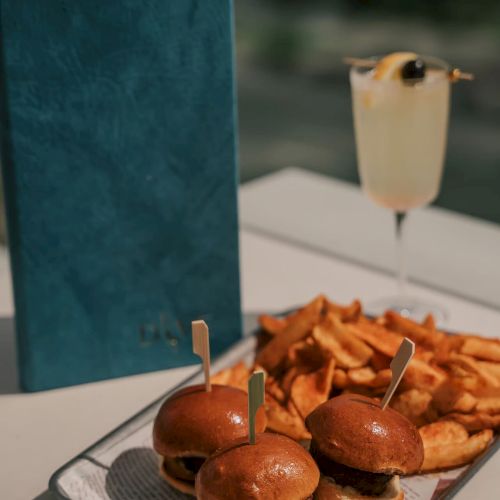 A tray with mini burgers and fries, a drink with a garnish, and a blue menu on a table.