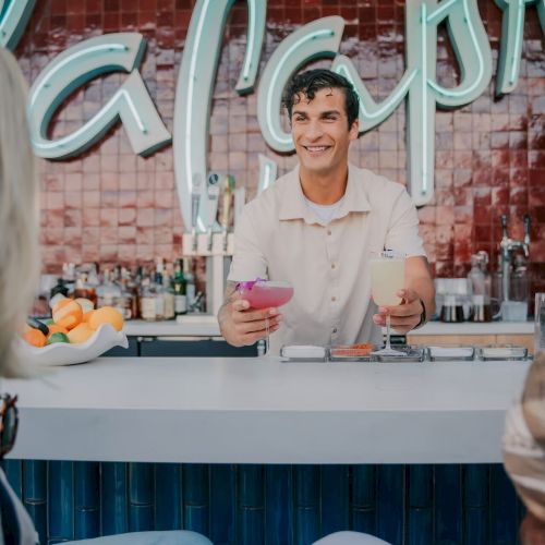 A bartender serves a pink drink at a bar with a neon sign and red tiled backdrop, while patrons sit at the counter.