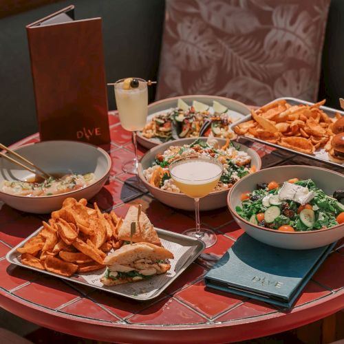 A table with various dishes: sweet potato fries, salads, drinks, and a sandwich. A menu and napkins are also present.