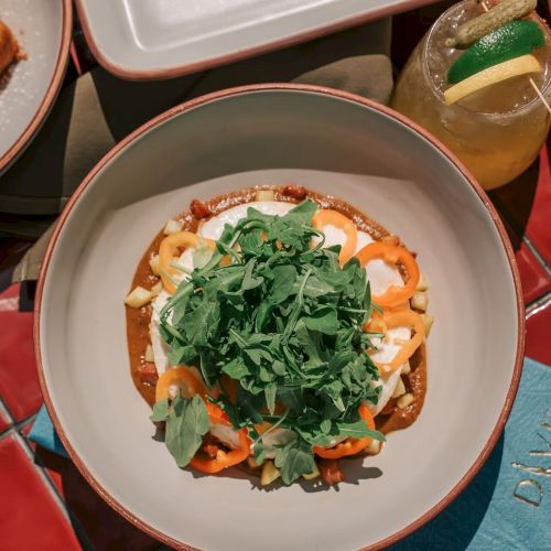 A plate of food topped with arugula and sliced vegetables, with a drink garnished with a lime wedge nearby on a colorful tile surface.