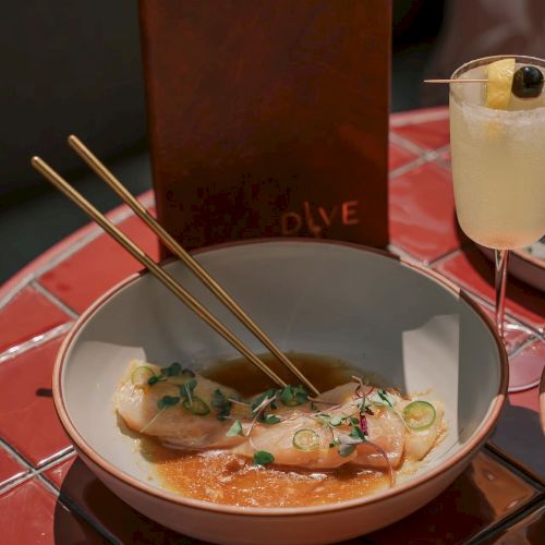 A plate with food served with sauce and herbs, chopsticks resting on top. A drink with a garnish and a menu are also present.