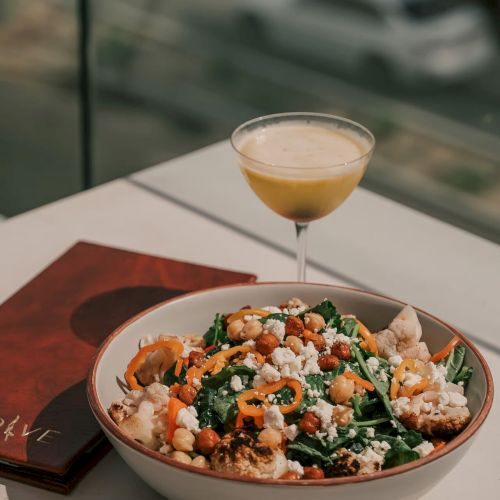A bowl of salad with various ingredients and a drink on the side sit on a table, with a menu placed nearby under sunlight.
