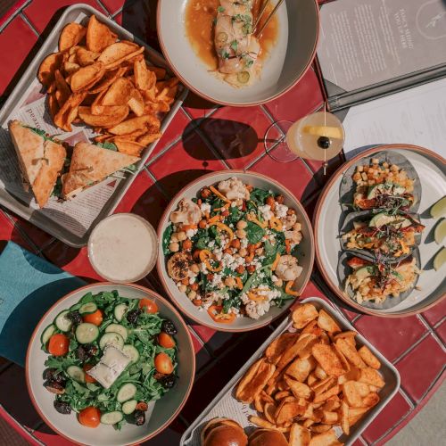 The image shows a table with various dishes: salads, sandwiches with chips, rolls, and drinks on a red surface.