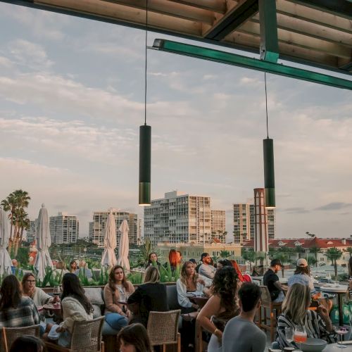 Outdoor dining area with people socializing, overlooking a cityscape and water, under a partly cloudy sky.