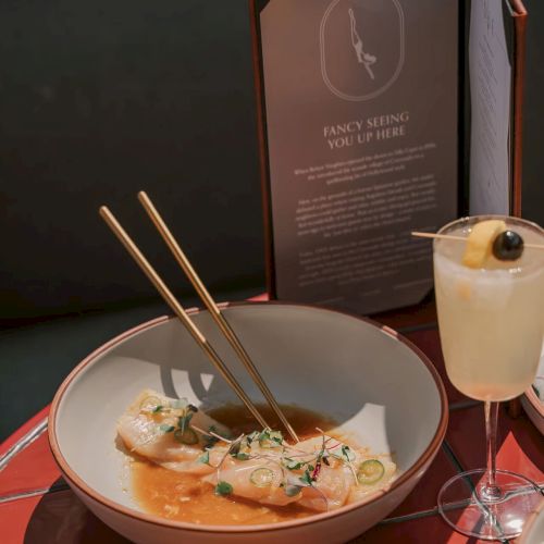 A bowl of food with chopsticks, a cocktail with a cherry, a menu, and a dish with orange-colored food on a red table.