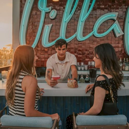 Two people sit at a bar with drinks, facing a bartender. A neon sign reading "Villa" is in the background.