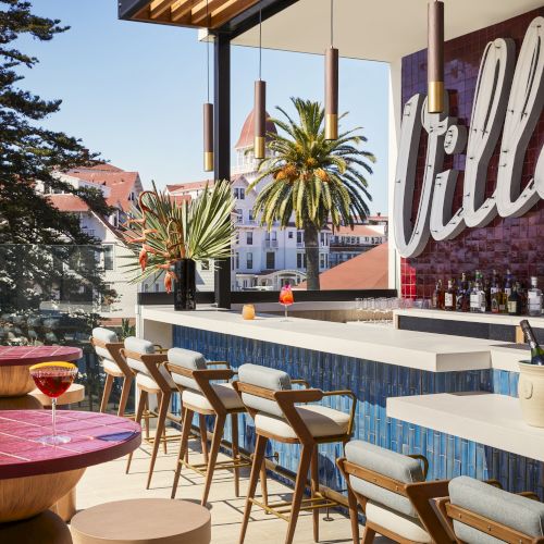 Rooftop bar with modern chairs, tables, and a bar area, overlooking palm trees and a cityscape, in a sunny setting.